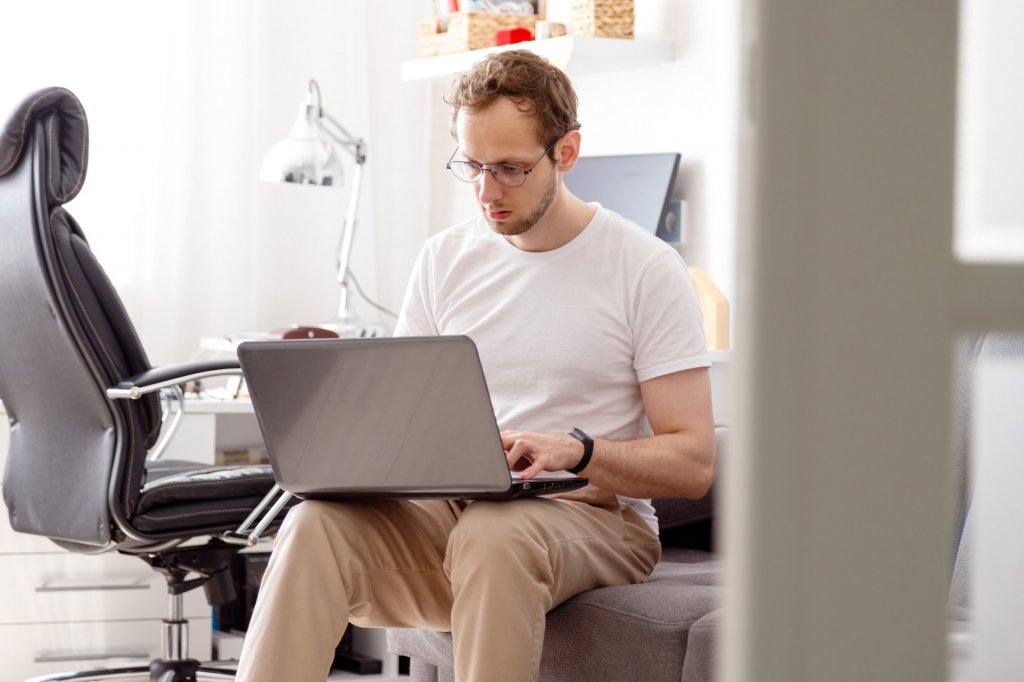 Young man with beard, working at home in living room sitting on coach with laptop